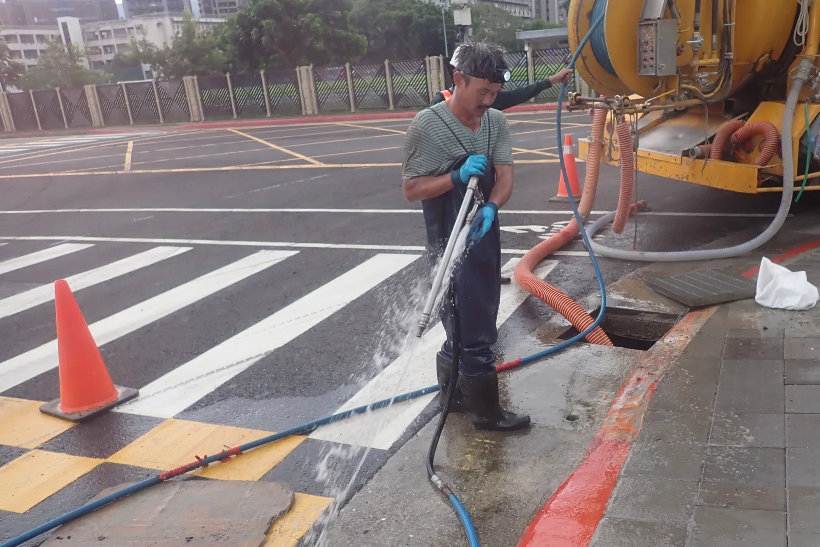 斑馬線旁的水溝口,師傅拿高壓水槍沖洗,後面是黃色清溝車與三角錐