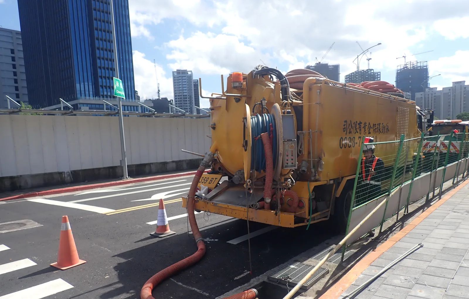 黃色清溝車停在新建大樓群旁的路邊，吸管一路拉到路緣掀開的溝口，車後擺著三角錐