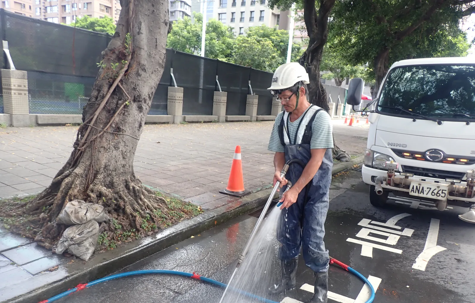 白帽師傅用水管沖洗路面，旁邊是行道樹與水車