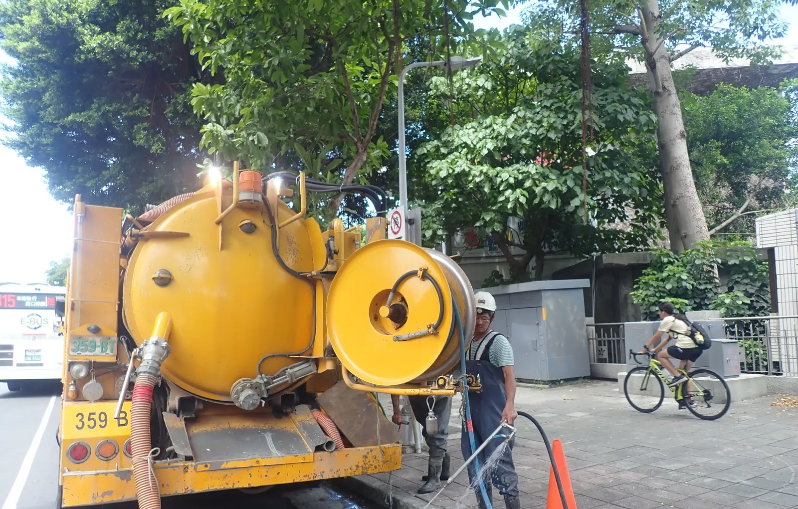黃色清溝車停在大樹下作業,後方是整排綠樹