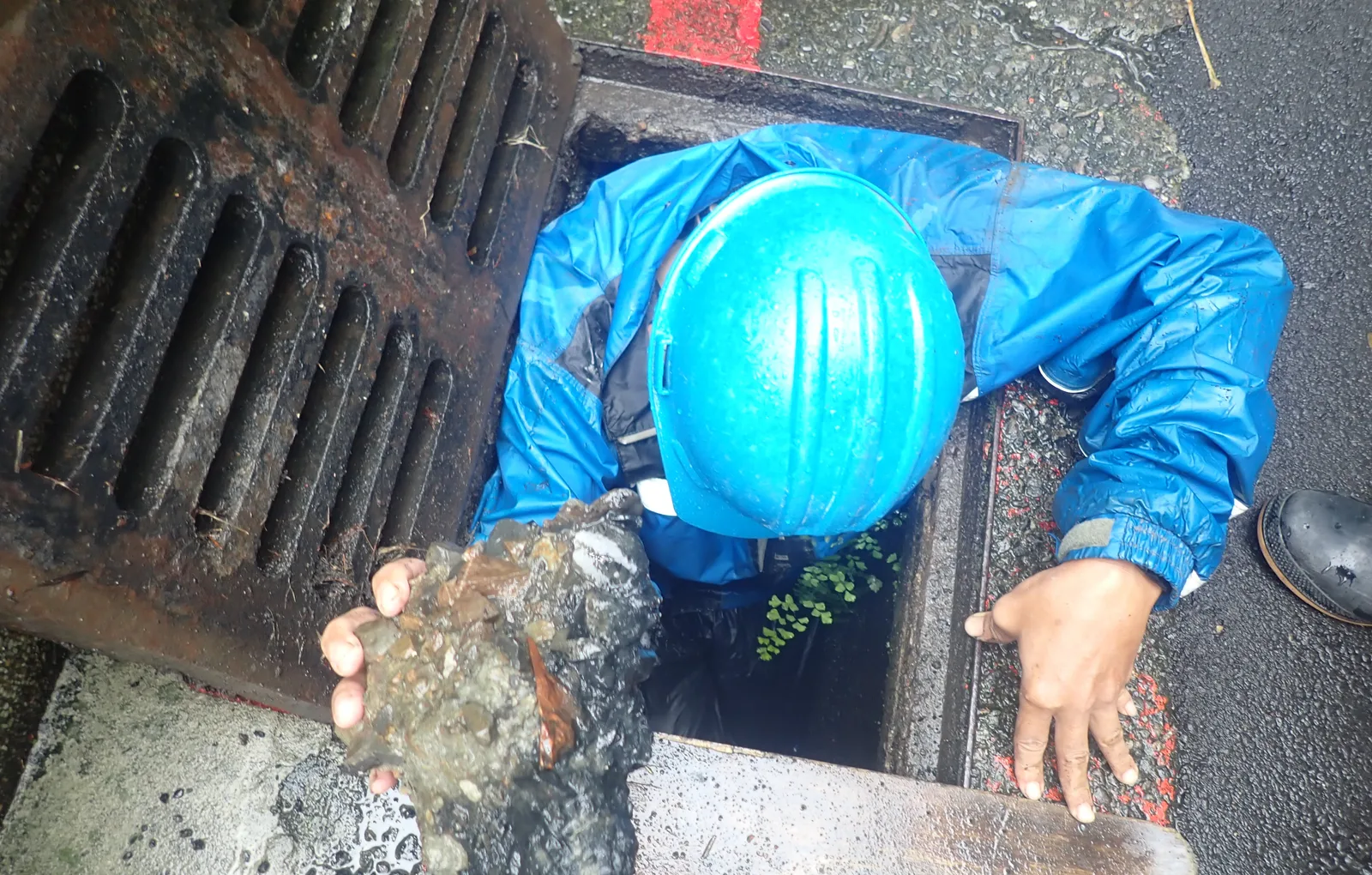穿藍色雨衣、戴藍色安全帽的師傅從格柵下的溝口撈起一大塊淤泥